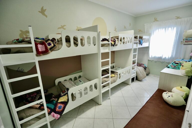 dormitorio-infantil-tres-beliches-vista-geral - ambiente com estilo Vista geral do dormitório infantil com três beliches brancos, mantas de crochê, almofadas e janela com cortina clara.
