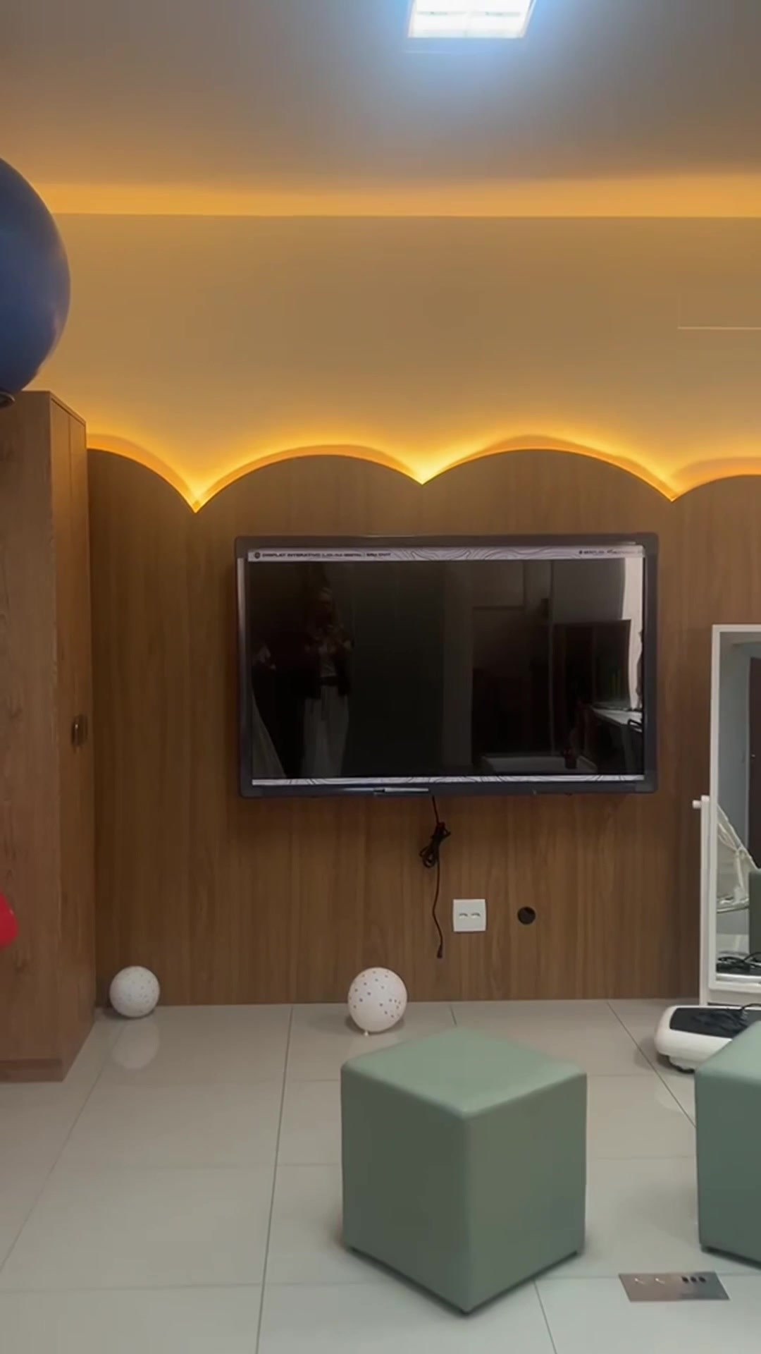 Painel ondulado em madeira com iluminação embutida no rebaixo, televisão e pufes verdes, na sala executada