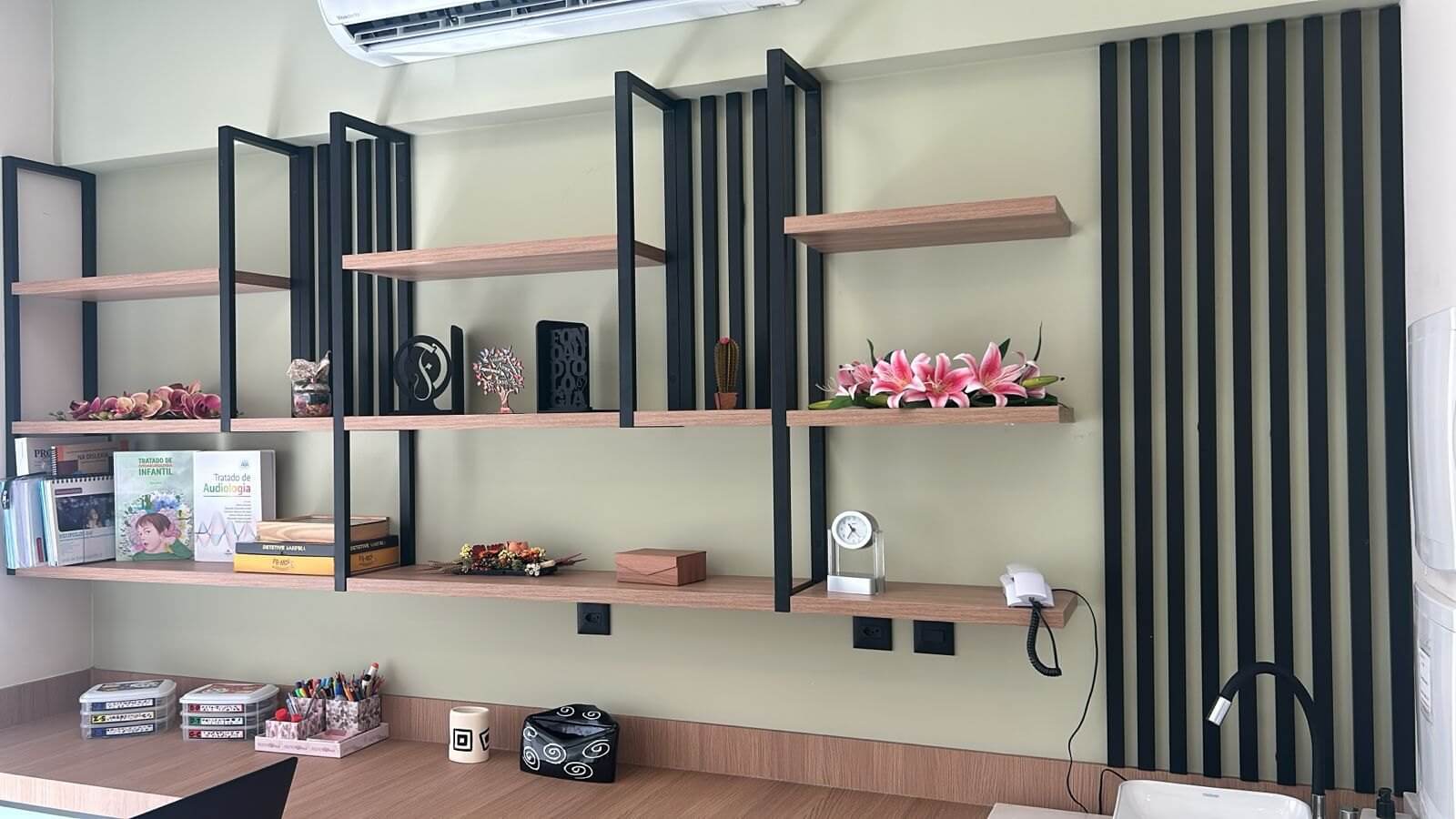 Prateleiras de madeira clara sobre estrutura metálica preta, fixadas em parede verde-sálvia com painel ripado ao lado e bancada de madeira abaixo.